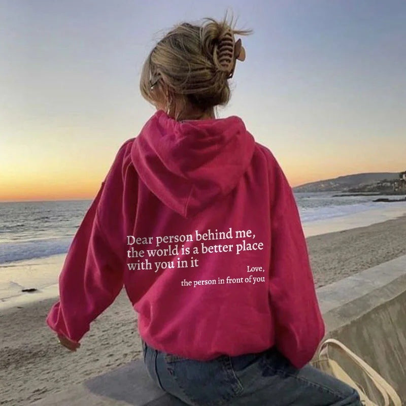 printed hoodie with pockets in pink worn by woman by the beach, featuring positive text print on the back.